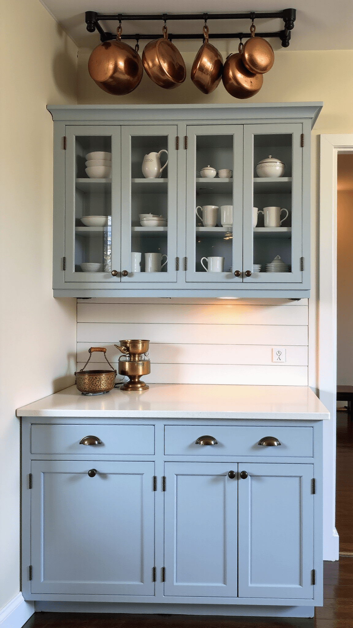 Intimate kitchen corner with blue-gray cabinets displaying white ironstone collection, copper pots hanging from ceiling rack, white shiplap backsplash, and vintage ceramic knobs, bathed in soft afternoon light.