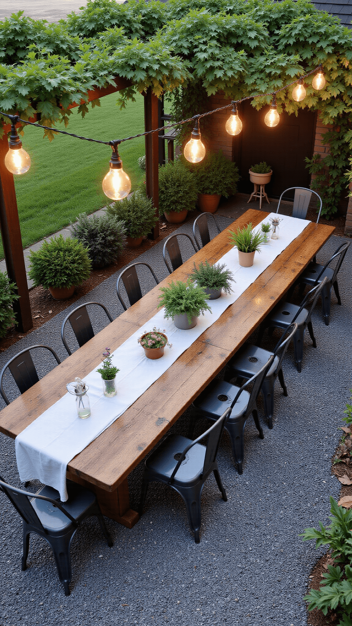 Bird's eye view of a rustic outdoor dining setup in late afternoon light, featuring a long, reclaimed wood farm table with a linen runner, vintage metal chairs on slate grey gravel patio, all under a pergola draped with grapevines amidst potted herbs and flowering plants, and enhanced by warm string lights.