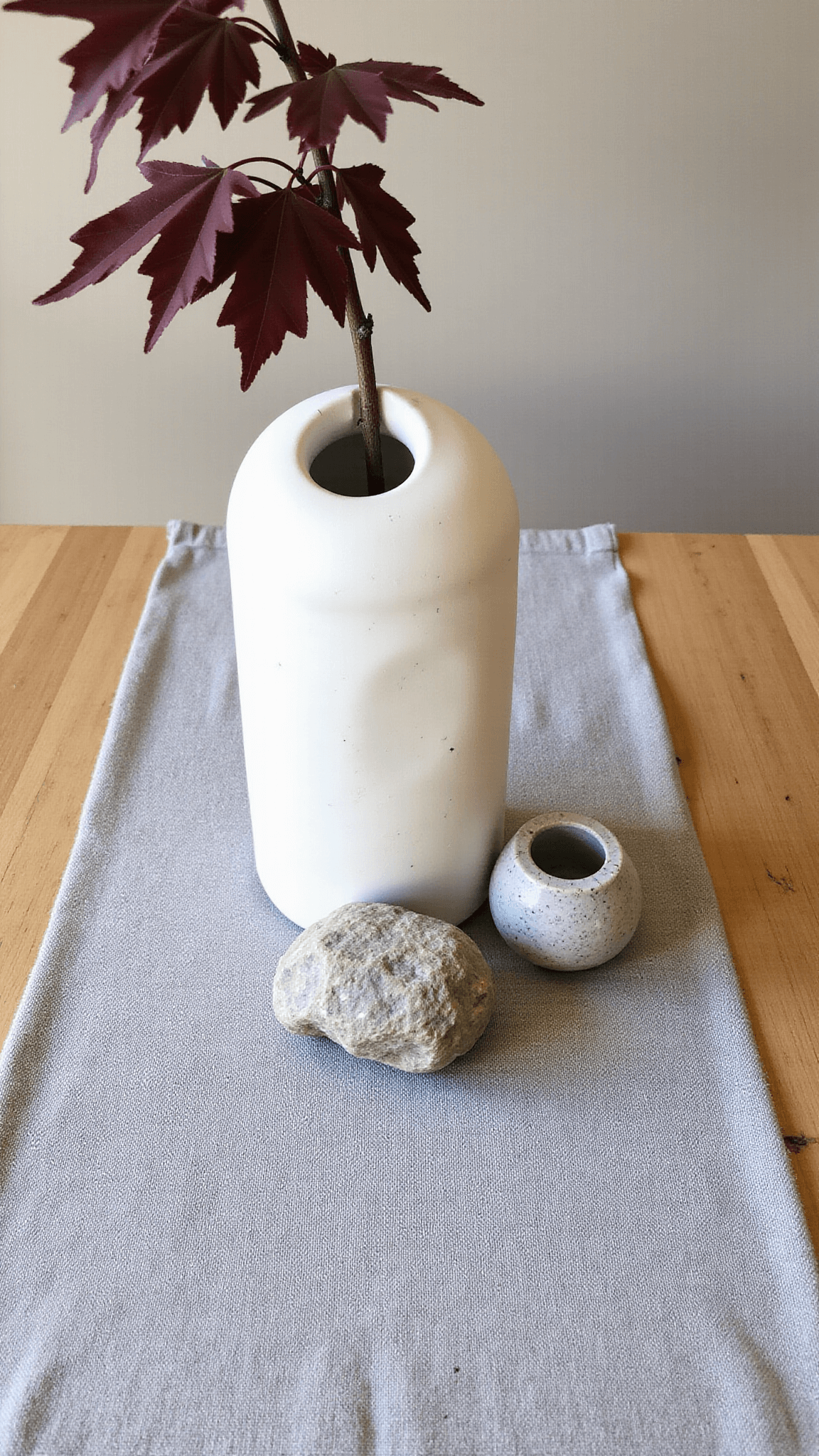 Overhead view of a styled dining table with a white ceramic vessel holding a Japanese maple branch, flanked by two stone vessels on a grey linen runner, in diffused midday light.