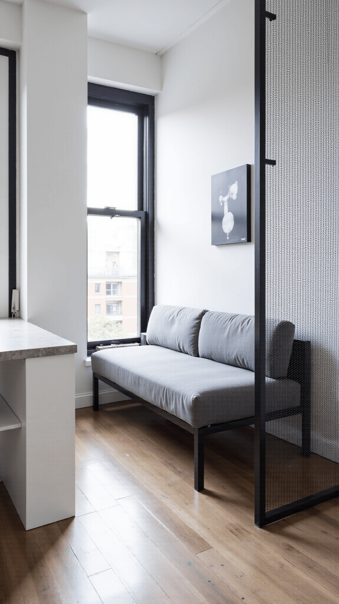 Minimalist city apartment with high-gloss white walls, black steel-framed windows, Murphy bed system, concrete side tables, low-profile gray sofa, and a wire mesh room divider featuring a monochromatic geometric art installation, all bathed in cool morning light.
